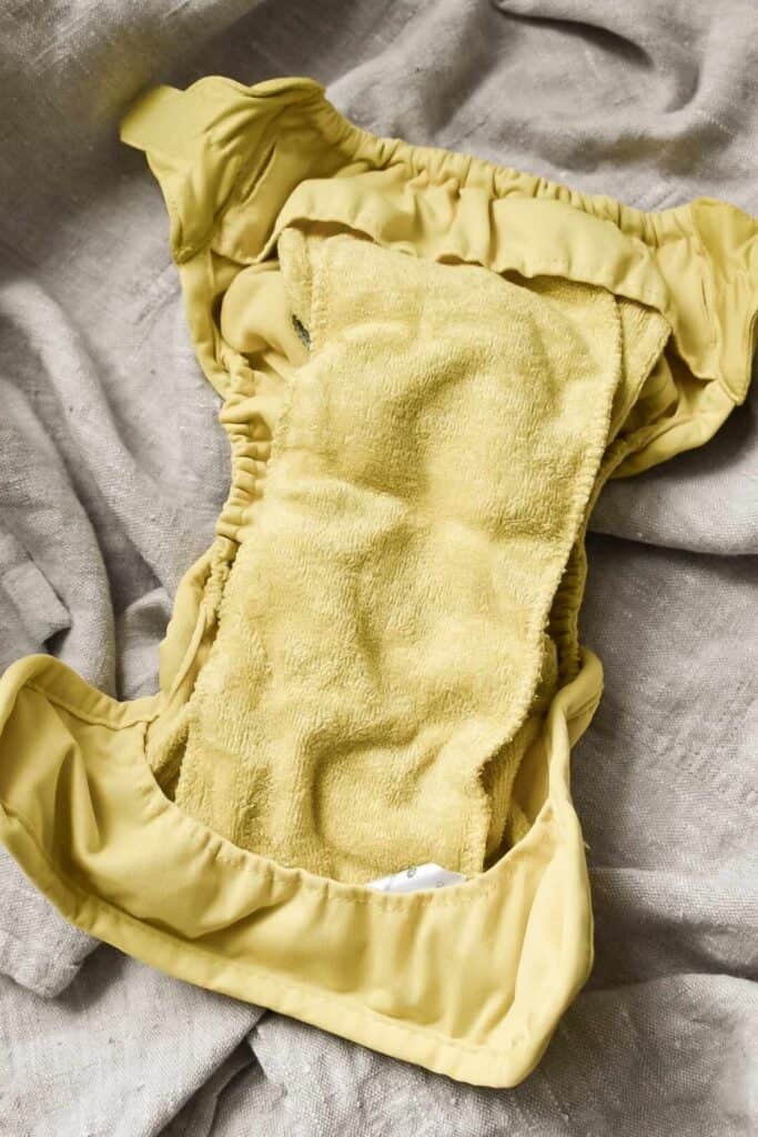 A reusable cloth nappy with a soft, yellow inner lining is laid open on a textured beige fabric surface. The diapers elastic edges are visible, showcasing its shape and structure—offering insights into different types available for your parental guide.