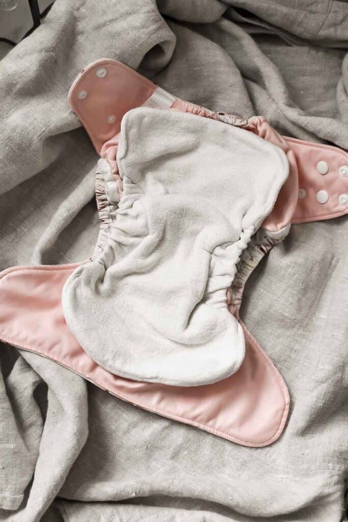 A pink cloth nappy with white inner lining is laid out on a textured gray fabric surface. This type features snap buttons for fastening and an elasticized waist, offering both comfort and practicality.