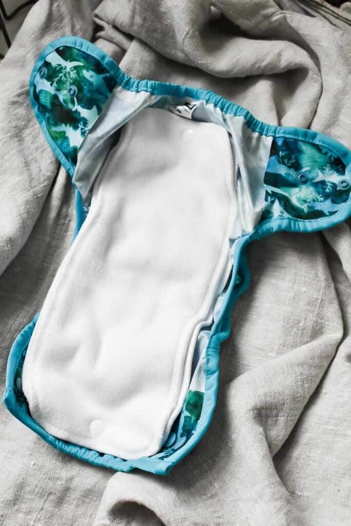 A cloth nappy with a white absorbent interior and a blue, forest-patterned exterior rests on a textured grey fabric surface.