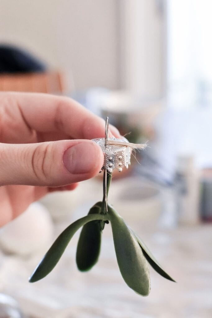 A hand holds mistletoe stuck to the cap of a glass bauble.