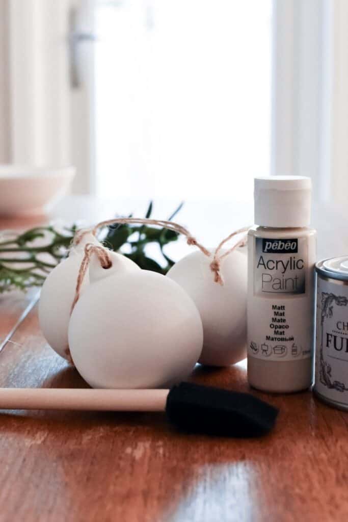 White ceramic ornaments with twine, a bottle of acrylic paint, a foam brush, greenery, and a tin of wax rest on a wooden table.
