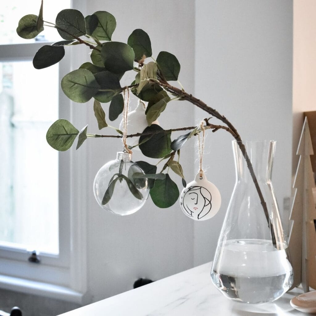 A clear vase with water holds a branch adorned with green leaves. Three DIY baubles hang from it: a transparent globe with a leaf inside, a painted bauble and a round white ornament featuring a minimalist face drawing.