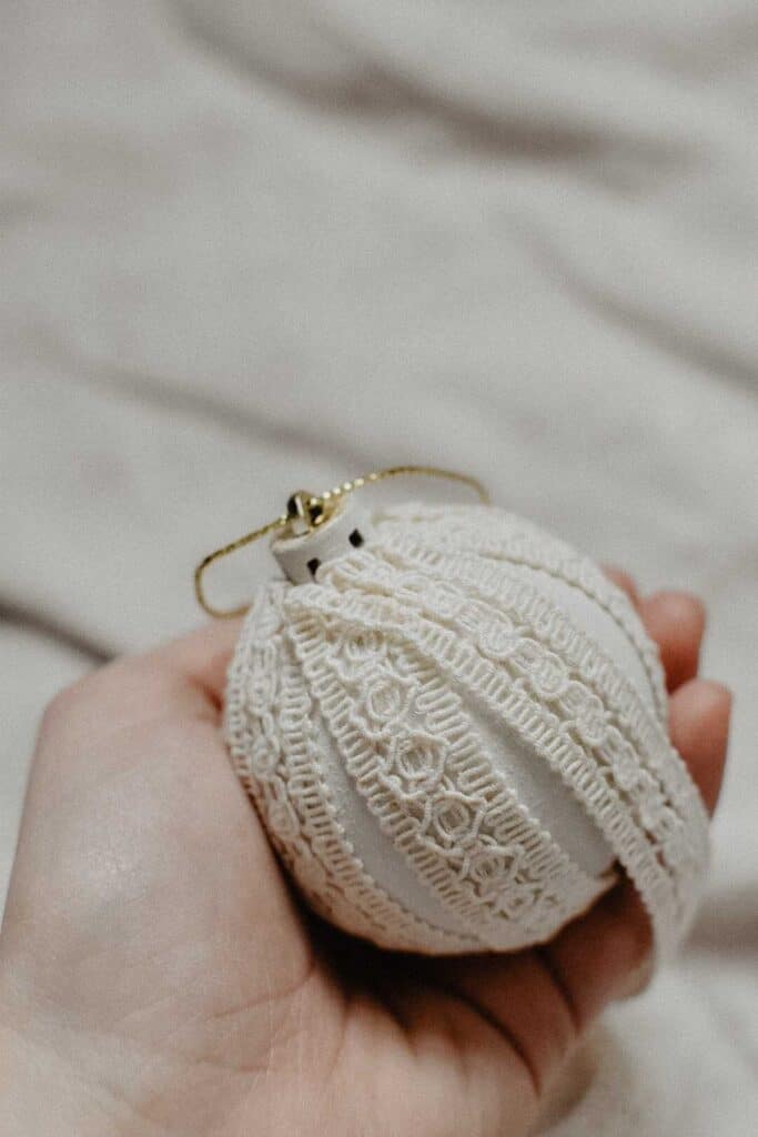 A hand holds a white decorative Christmas ornament crafted from recycled materials, featuring intricate lace patterns and a gold loop. The background is a softly blurred, neutral-colored fabric.