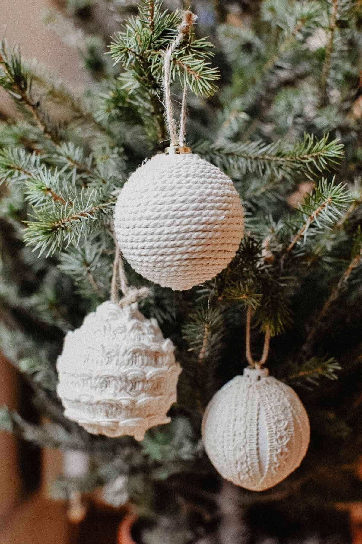 Three white, upcycled Christmas decorations hang from a green Christmas tree branch. Crafted from recycled materials, each ornament boasts a unique pattern, adding a cozy, upcycled touch to the festive decoration.