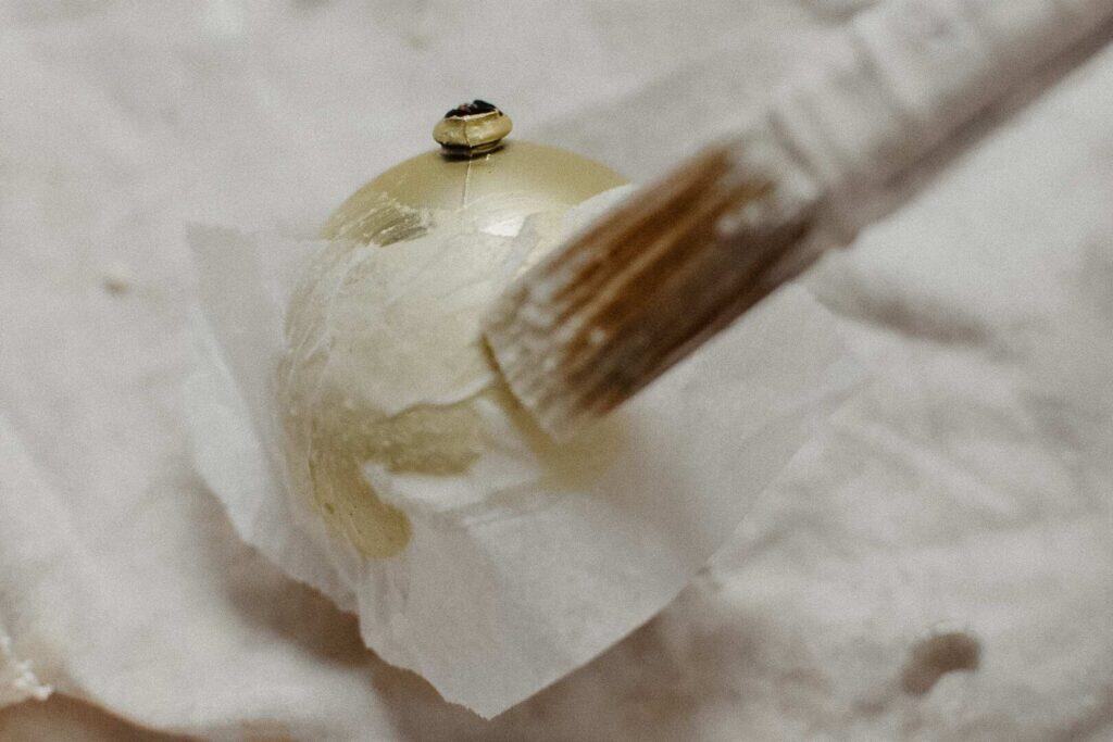 A paintbrush applies white paint to a gold Christmas ornament, transforming it into a stunning upcycled Christmas decoration resting on crumpled white paper.