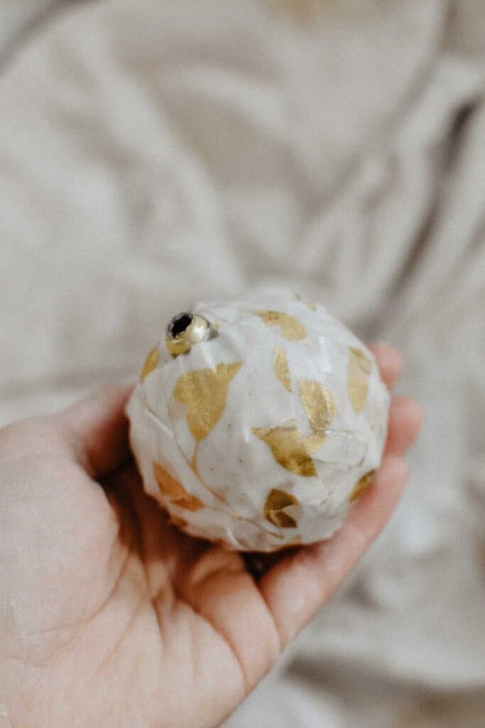 A hand holds a decorative spherical object with a white surface and gold leaf patterns, reminiscent of elegant Christmas decorations. The background is softly blurred, highlighting the objects beauty and festive charm.