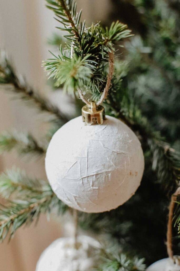 Close-up of a white, textured bauble crafted from recycled materials hanging on a green pine branch. The decoration is attached with a brown string, and the background shows blurred pine needles, suggesting an upcycled Christmas setting.