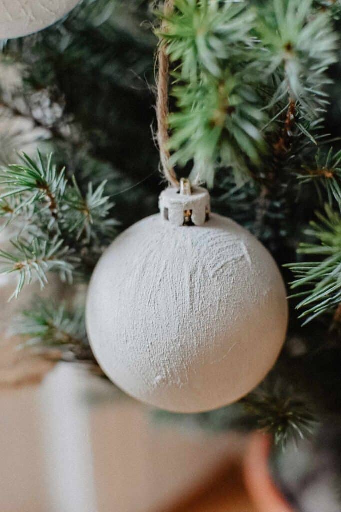 A close-up of a white textured Christmas ornament hanging from a green artificial tree branch. This bauble, crafted with recycled materials, has a rustic appearance and is attached using twine, adding a simple and natural charm to the Christmas decorations.