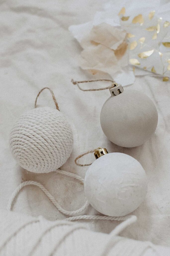 Three decorative Christmas ornaments on a soft beige surface crafted from recycled materials. One ball is textured with rope, another smooth and matte, while the third boasts a slightly glossy finish. A piece of white and gold wrapping paper peeks from the background.