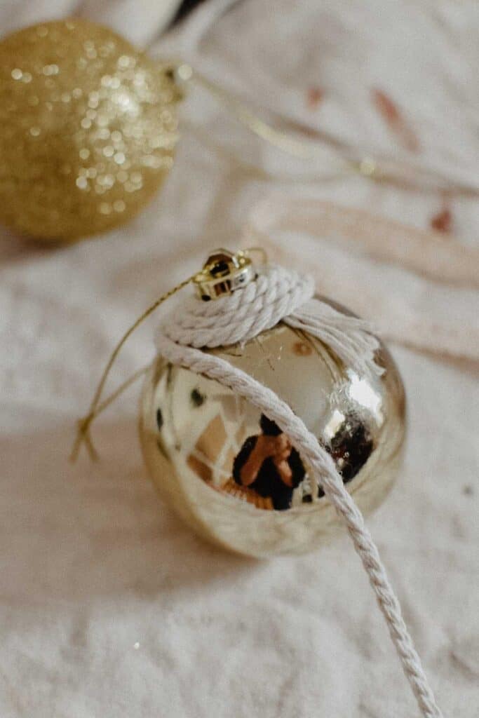 A shiny gold Christmas ornament, crafted from upcycled materials, lies on a white surface reflecting nearby objects. It is partially wrapped with white cord. Another blurred, glittery gold ornament from recycled decorations shimmers softly in the background.