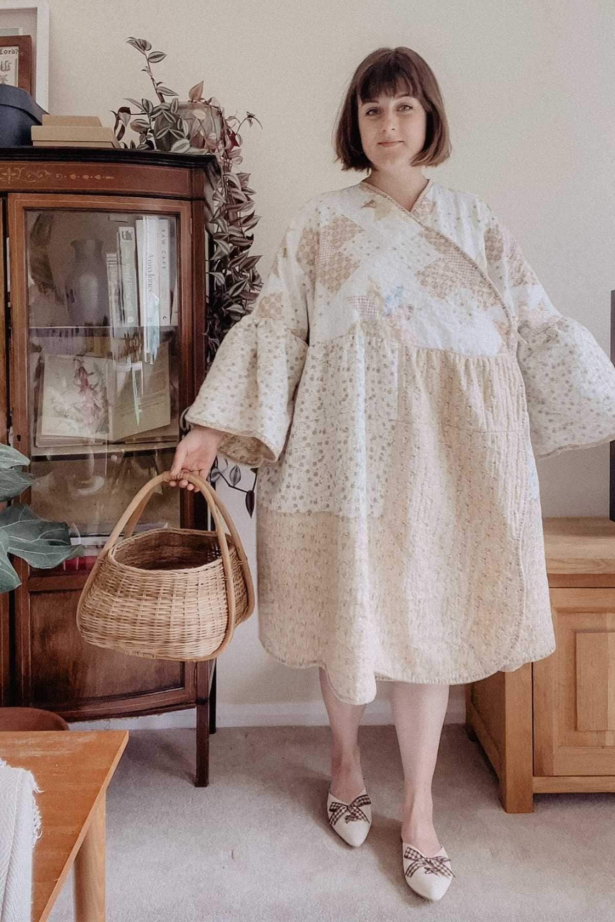 A person wearing an upcycled dress crafted from old quilts stands indoors holding a wicker basket. She is in a room with a wooden cabinet, potted plant, and light wood furniture. The setting feels cozy and homey, reflecting the charm of upcycling fashion.