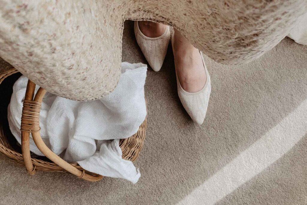 Overhead view of a person in cream-colored woven VIVAIA flats standing on a carpet, wearing a textured beige dress made from old quilts. Next to them is a wicker basket with a white cloth inside.