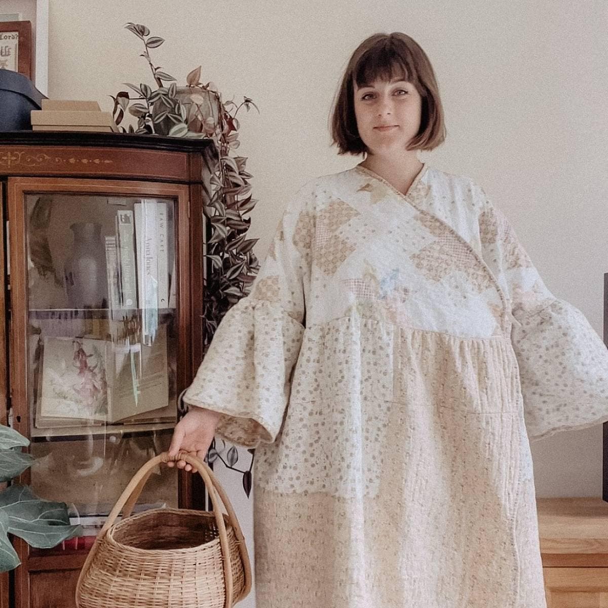 A person stands indoors holding a wicker basket, wearing a long, upcycled dress made from old quilts with wide sleeves. They are in front of a wooden cabinet adorned with books and a plant. To the left, a large leafy plant peeks into view.