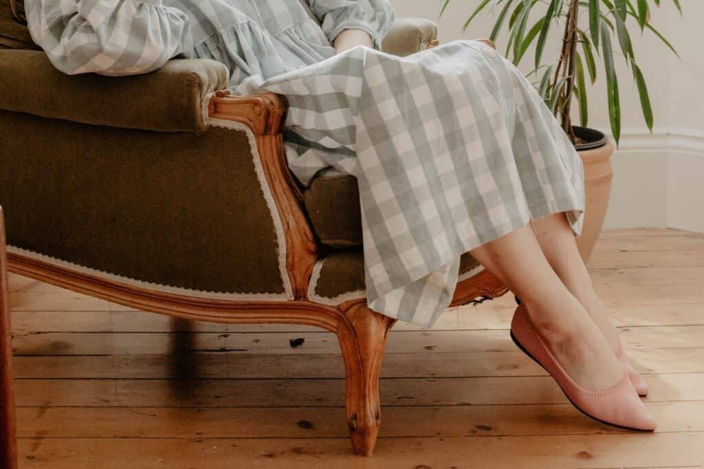 A person in a checkered dress sits in a vintage armchair with wooden trim, their feet adorned with pink sustainable shoes by VIVAIA. A potted plant adds to the cozy and tranquil setting.