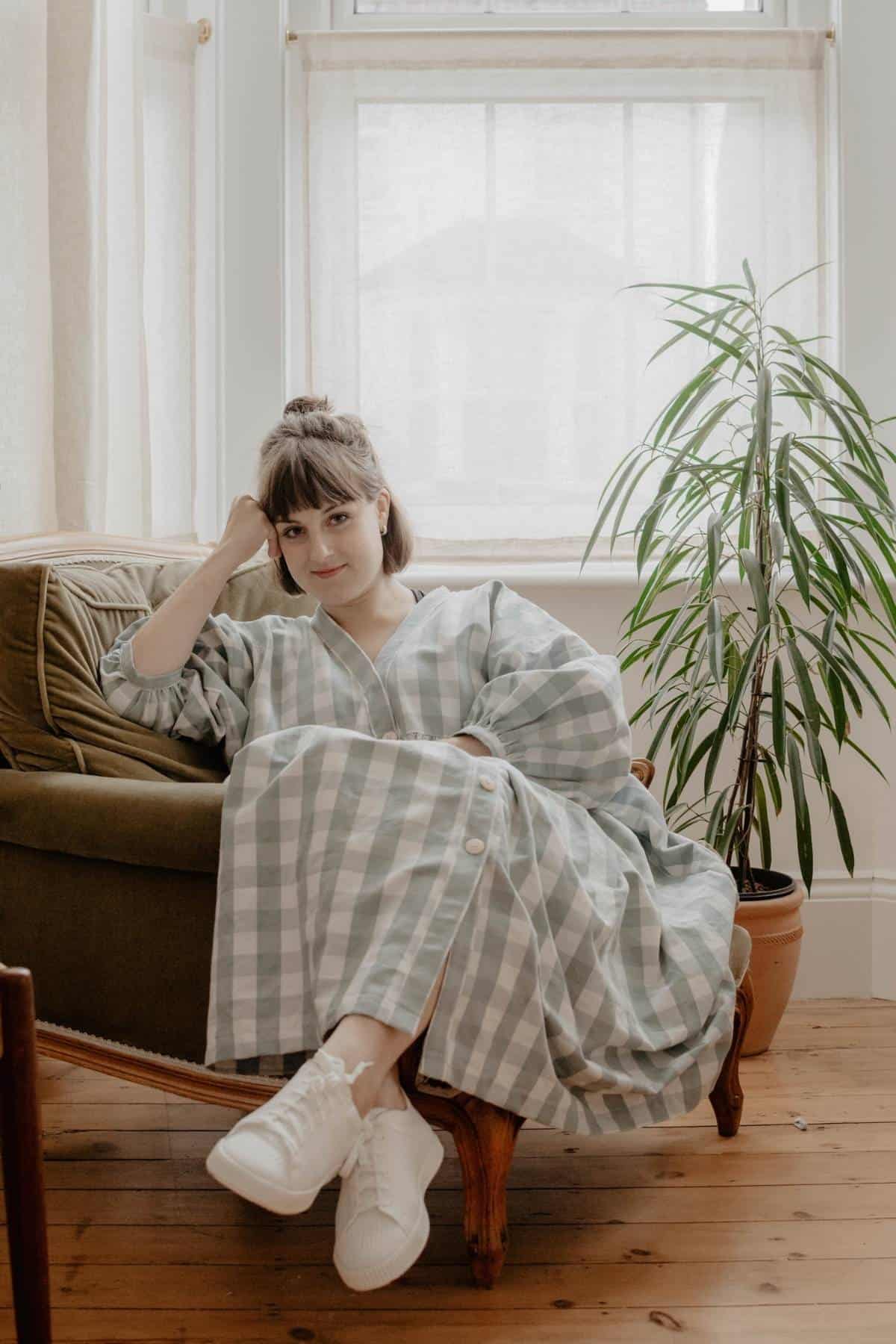 A person with brown hair tied up, wearing a long checkered dress and white VIVAIA sneakers, sits on a sofa. They rest their head on their hand, looking relaxed. A tall plant is beside them, and light filters through sheer curtains, creating a serene scene.