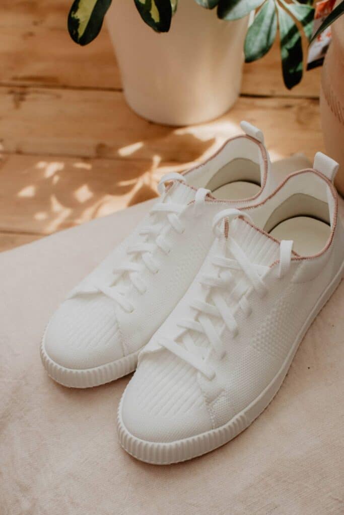 A pair of VIVAIA white sneakers with laces rests on a light-colored surface, bathed in soft natural light. A plant with green leaves partially peeks into the background, adding a fresh touch to this scene of sustainable shoes.