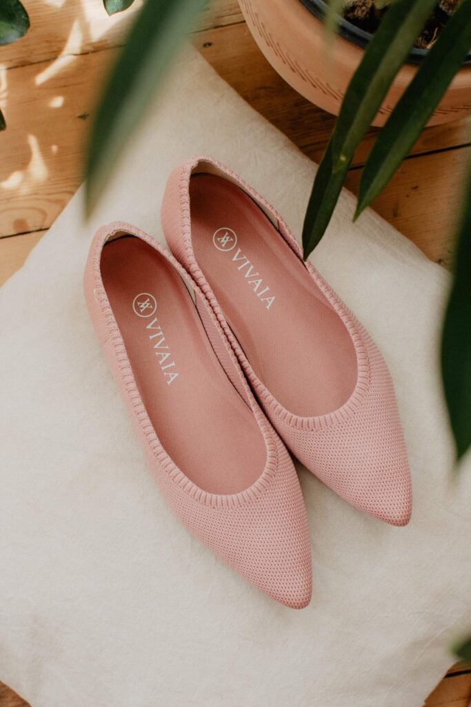 A pair of pink knitted flats from VIVAIA with pointed toes are placed on a white cloth. These sustainable shoes have a subtle texture, with VIVALA written on the insole. Green leaves partially frame the image, offering an elegant touch to this eco-friendly design.