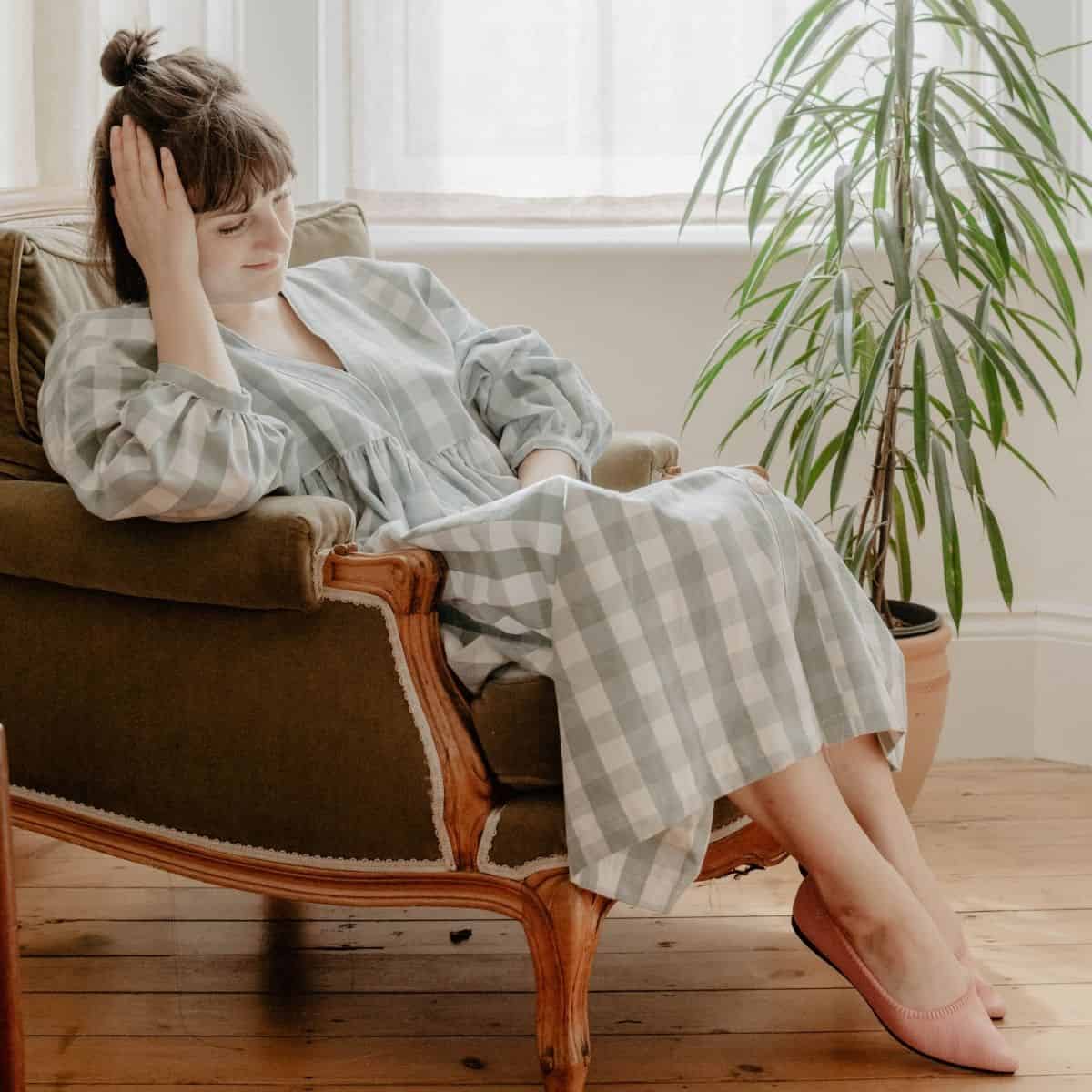 A woman in a checkered dress sits in an armchair with her eyes closed and head resting on her hand, as a potted plant thrives beside her. Sunlight filters through the window, highlighting her VIVAIA sustainable shoes, creating a calm ambiance.