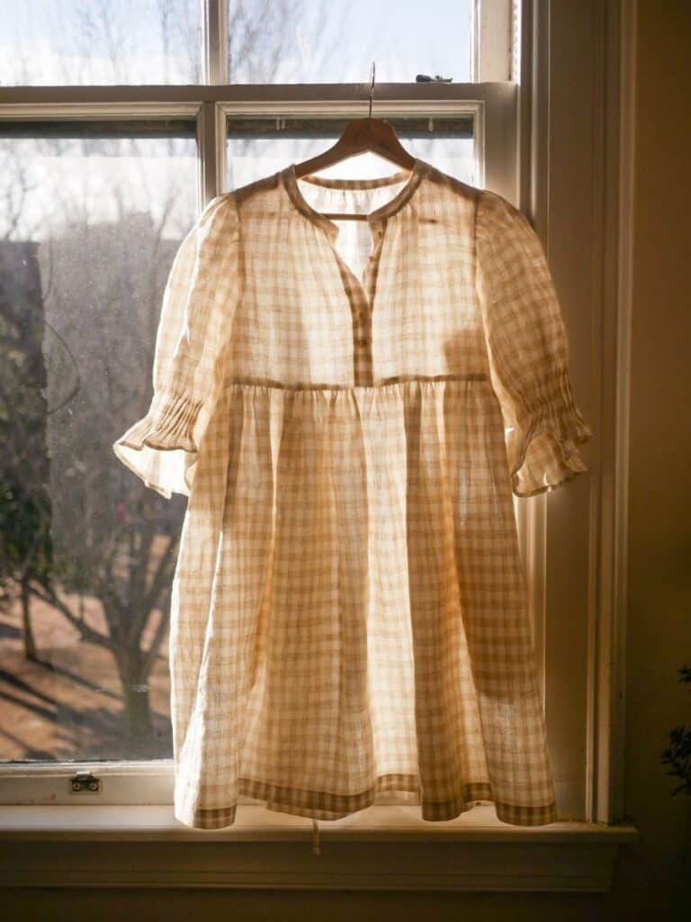 A soft, gauzy Vivien Shao Chen Orchards Dress hanging in front of a sunlit window, made from a light, gingham fabric with a loose, dreamy silhouette, embodying effortless cottagecore charm.