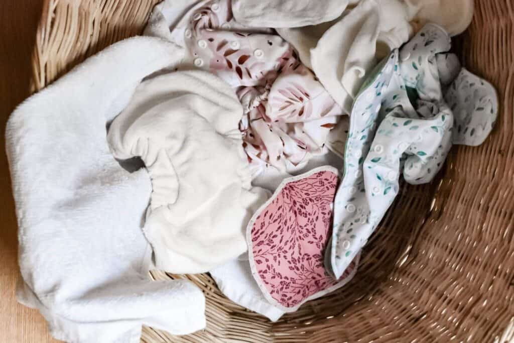 A wicker washing basket brimming with cloth nappies and nappy inserts showcases an array of patterns and colors. The mix includes whites, pinks adorned with leaves, and teal featuring abstract designs.