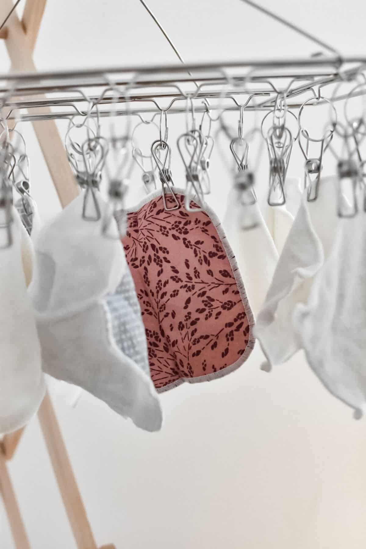 Freshly washed cloth nappy inserts and reusable wipes hang on a metal drying rack with clips. One pad boasts a pink and brown floral pattern.
