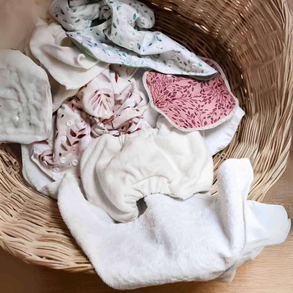 A wicker basket brimming with cloth nappies displays a kaleidoscope of colors and patterns, from white to pink floral prints and light green leaf designs. Perfect for those following a nappy washing guide, the basket rests charmingly on the wooden floor.