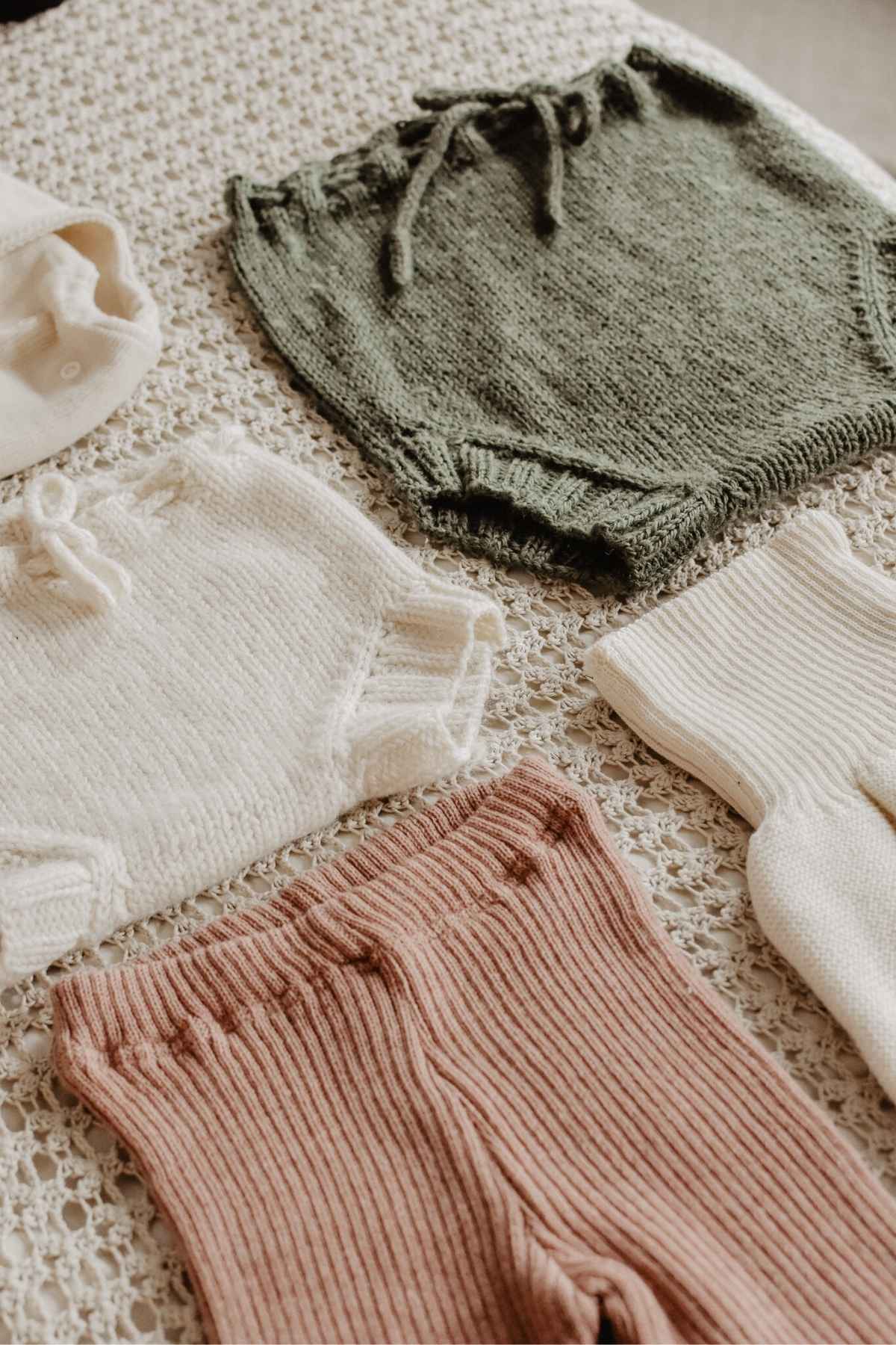 Assorted knitted baby clothing, including white and green shorts, a white sweater, pink and cream pants, and cozy wool nappy covers artfully arranged on a textured beige surface.