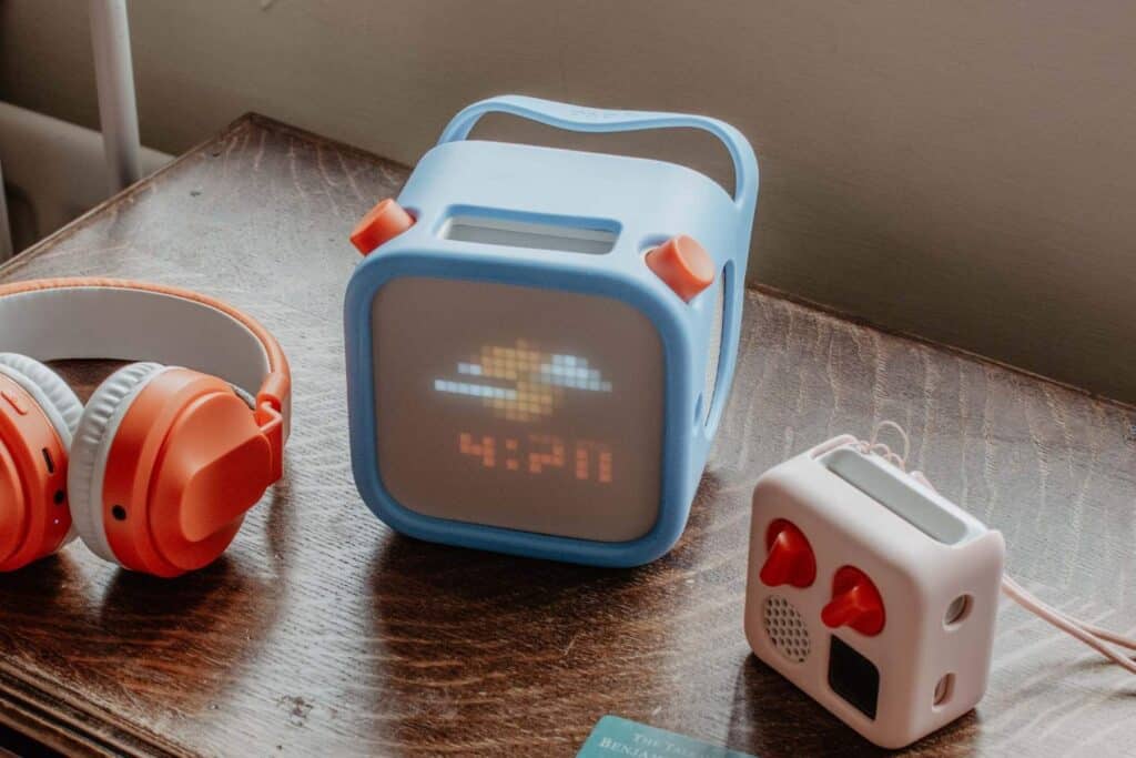 A blue Yoto player 3rd generation with pixelated cloud display shows 4:20, next to a small Yoto Mini with red knobs, and orange and white headphones, all on a wooden surface.