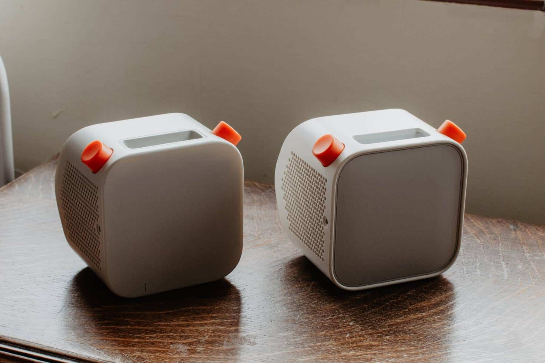 Two Yoto players with orange knobs  sit on a wooden table. The 2nd generation Yoto on the left is slightly larger than the 3rd generation Yoto player on the right. The speakers have a perforated side panel and a minimalist design, set against a neutral background.