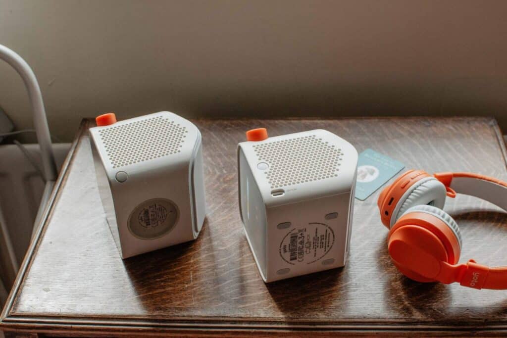 Two white Yoto players with orange accents are placed on their sides on a wooden surface beside orange and white headphones. Theres a blurred card in the background. The 2nd generation Yoto player on the left is very similar to the 3rd generation Yoto player on the right, except that the buttons and charging port are in slightly different places.