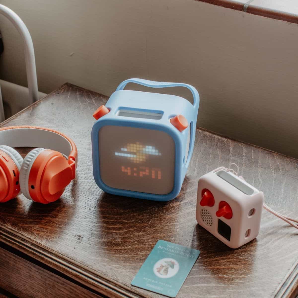 A wooden table with orange headphones, a 3rd generation Yoto player displaying pixel art and the time, a pink Yoto Mini, and a card of Benjamin Bunny.