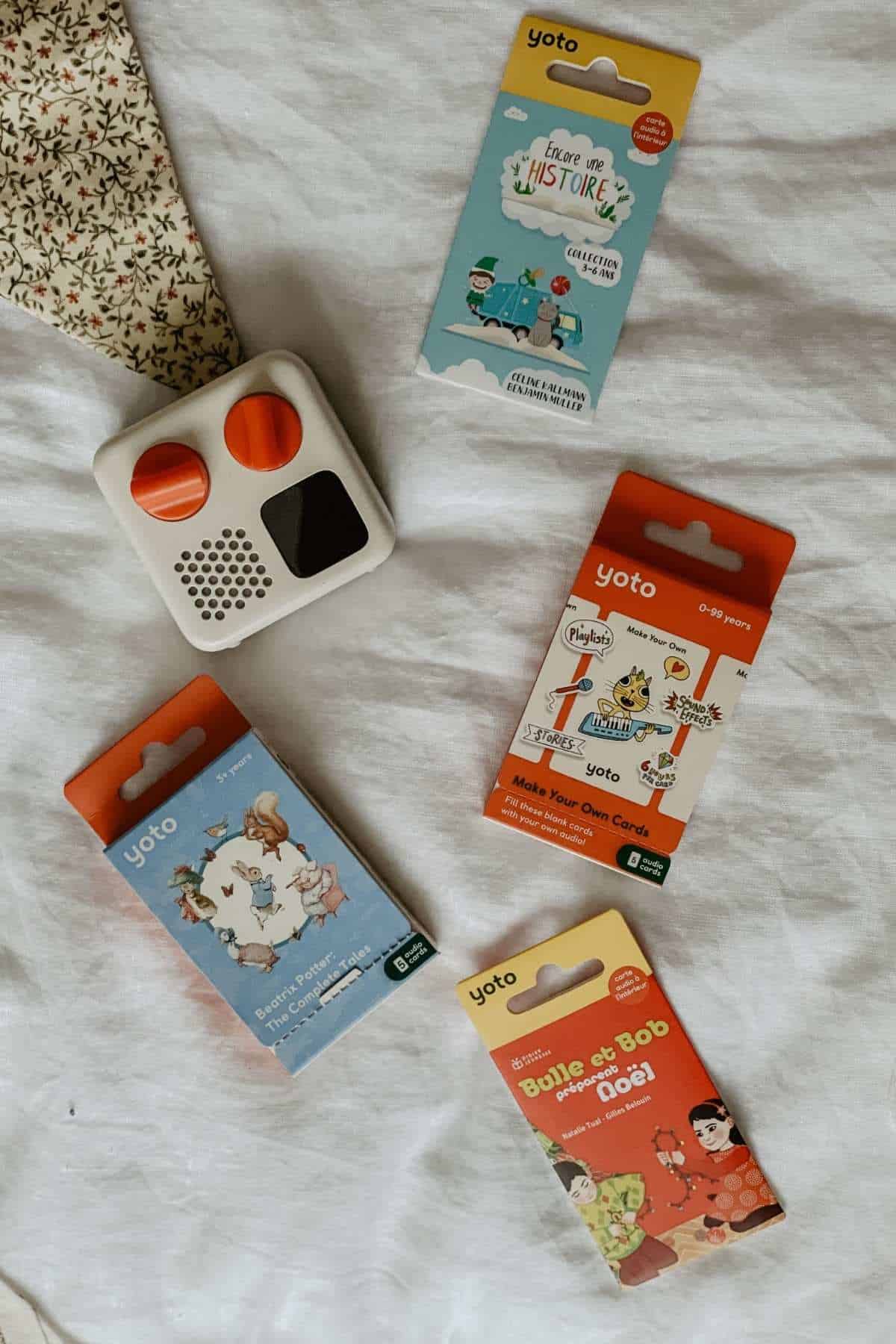 A small white and gray kids Yoto Mini audio player with round buttons sits on a white fabric. Surrounding it are five colorful card packs with playful illustrations and the brand name Yoto. Theres a patterned bunting in the top left corner.