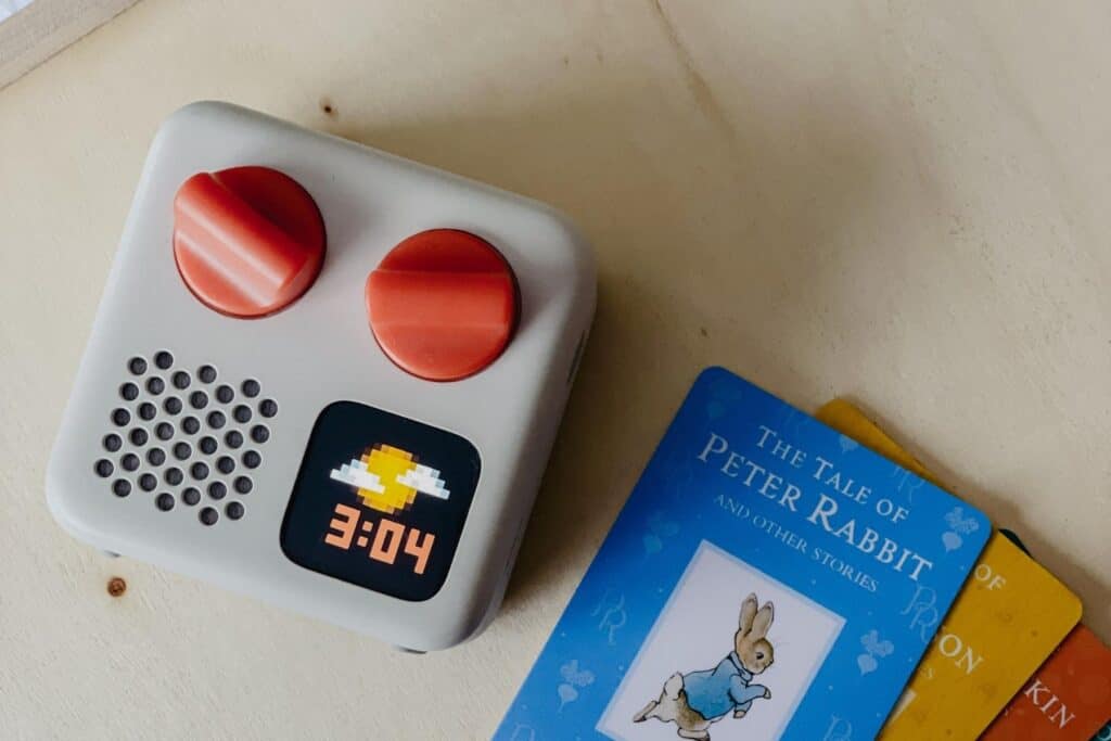 The white Yoto Mini kids audio player boasts two red dials and a small digital screen showing an icon and 3:04. Nestled beside it is The Tale of Peter Rabbit book and two vibrant cards, offering endless audio adventure for little listeners.
