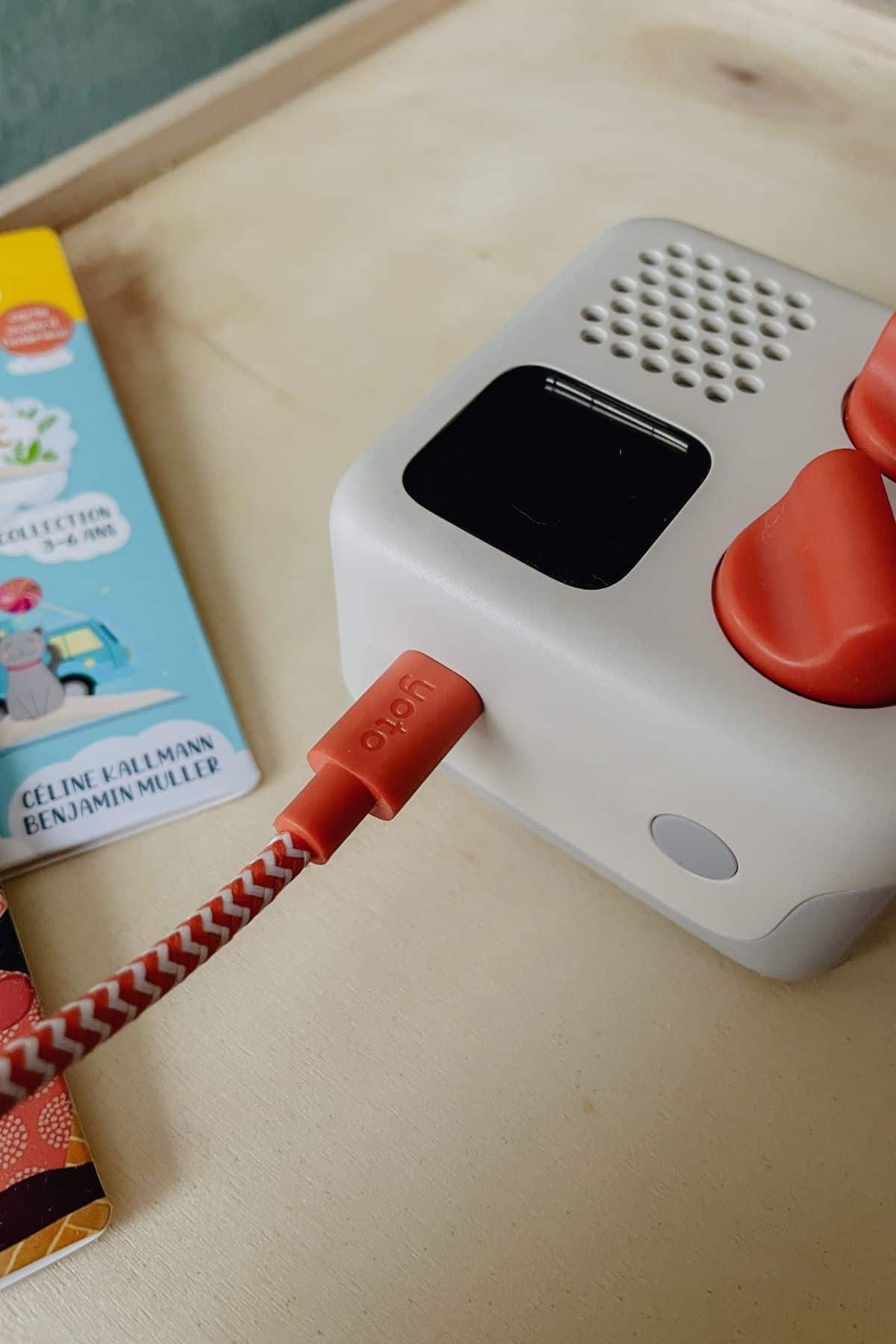 Close-up of a Toniebox, a gray kids audio player with a red figure on top, connected to a red charging cable. A childrens book with colorful illustrations is partially visible on the left, making it worth buying for little listeners. The setup rests on a wooden surface.