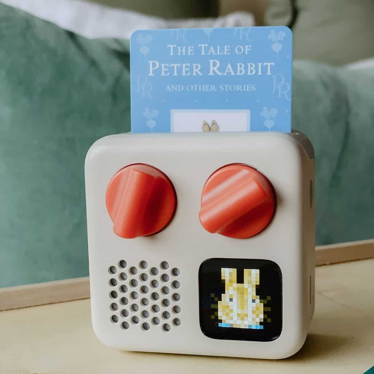 A small beige kids Yoto Mini audio player with two orange knobs displays a pixelated bunny on its screen. A card titled The Tale of Peter Rabbit and Other Stories is inserted into the top. The Yoto Mini rests on a wooden surface with a green cushion in the background.